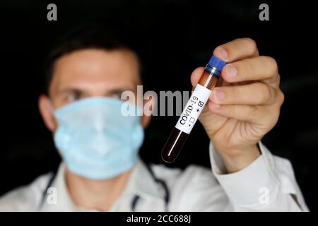 Provetta per analisi con campione di sangue covid-19 in primo piano con la mano maschio, uomo in maschera medica contenente un flaconcino. Medico o scienziato con test coronavirus positivo Foto Stock