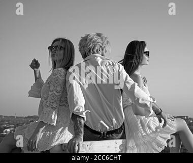 Due graziose ragazze e un uomo brutale che si siedono tra loro guardando in lontananza sullo sfondo del cielo azzurro Foto Stock