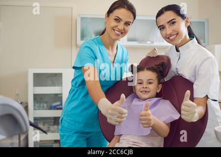 Dentista sorridente, assistente e bambina paziente mostrando pollici in su segno in clinica dentale Foto Stock