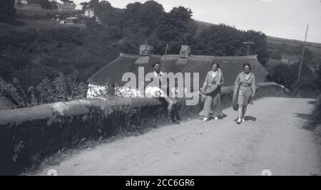 Anni '20, storico, 'andando alla spiaggia'....una madre e una figlia che camminano su un percorso polveroso su un ponte che porta grandi anelli di gomma, con il padre seduto sul lato del muro di mattoni che tiene una coperta, Devon, Inghilterra, Regno Unito. Un tetto tradizionale cottage con tetto in paglia può essere visto nella foto. Foto Stock