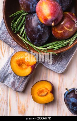 Frutta fresca di prugne dolce intera e tagliata in ciotola di legno marrone con foglie di rosmarino sul vecchio tagliere, sfondo del tavolo di legno, vista dall'alto Foto Stock