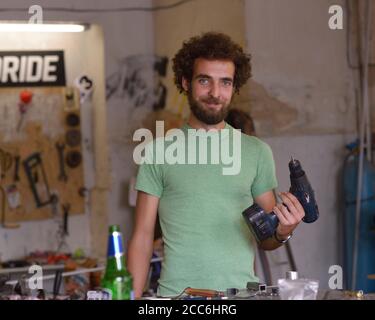 Giovane uomo di riparazione sorridente portandogli in piedi al tavolo da lavoro con un trapano in una mano presso il suo negozio di riparazione bici. 23 luglio 2019. Kiev, Ucraina Foto Stock