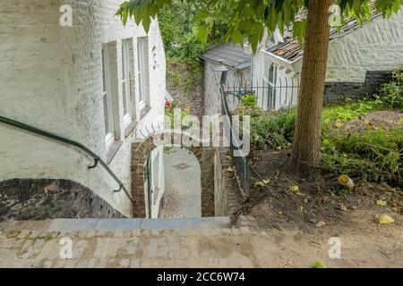 Prospettiva di un vicolo con una scala giù con un ringhiera metallica tra una casa con una parete bianca e. Un piccolo giardino con una parete ad arco in Maestri Foto Stock