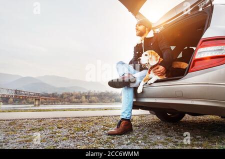 Uomo impanato in abiti caldi seduto con beagle nel bagagliaio dell'auto. Viaggiare con animali domestici Foto Stock