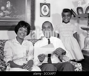 RITRATTO DEGLI ANNI '60 DELLA SORRIDENTE FAMIGLIA AFROAMERICANA DI 3 PERSONE SEDUTE COUCH PADRE CHE TIENE BUSTA CON CONTROLLARE TUTTI GUARDANDO LA MACCHINA FOTOGRAFICA - N1807 HAR001 CONTROLLO HARS PER INTERNI COPPIA NOSTALGICA SUBURBANA URBANA LE MADRI INVILUPPO VECCHIO TEMPO NOSTALGIA VECCHIA MODA 1 ASSICURAZIONE GIOVANILE DIVANO STILE DI COMUNICAZIONE SODDISFATTO FAMIGLIE GIOIA STILE DI VITA SODDISFAZIONE DONNE SPOSATE STUDIO SHOT CONIUGE HUSBAND HEALTHINESS HOME VITA COPIA SPAZIO AMICIZIA MEZZA LUNGHEZZA SIGNORE FIGLIE PERSONE MASCHI FIDUCIA PADRI B&W PARTNER OCCHIO CONTATTO FELICITÀ ALLEGRO AFROAMERICANI AFROAMERICANI PAPÀ ECCITAZIONE NERO ETNIA ORGOGLIO Foto Stock