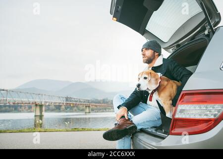 Uomo impanato in caldo abito casual autunno siting con beagle nel bagagliaio dell'auto. Viaggiare con animali domestici. Foto Stock
