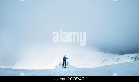 Inverno Trekker rimanendo sulla cima della montagna che ha scalato e. godendo di un'ampia vista panoramica della valle coperta da nubi tempeste Foto Stock