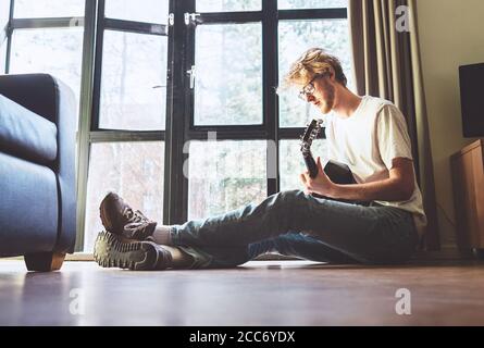 Middle shot di un giovane suona sulla chitarra seduto sul pavimento in soggiorno. Immagine del concetto di formazione musicale. Foto Stock