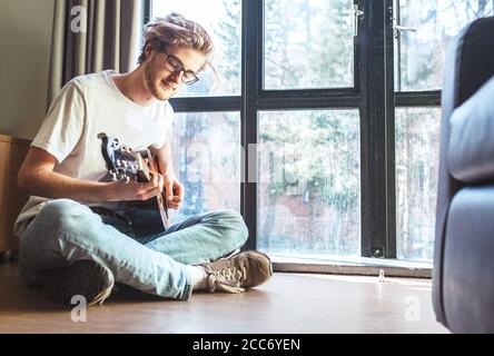 Middle shot di un giovane suona sulla chitarra seduto sul pavimento in soggiorno. Immagine del concetto di formazione musicale. Foto Stock