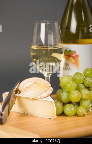 Baby Brie affumicato, uva, cracker d'acqua, coltello, bicchiere e bottiglia di vino chardonnay sul tagliere Foto Stock