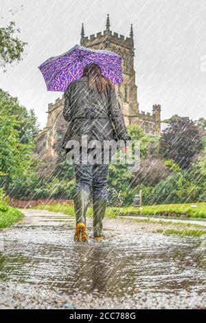 Vista posteriore di donna che indossa i wellies & che trasporta l'ombrello che cammina sotto la pioggia pesante isolato sul canale britannico alzaia. Foto Stock