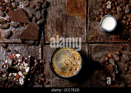 Tazza di caffè espresso con caraffa di latte, cioccolato fondente fatto a mano, caffè e chicchi di cacao intorno a piastrelle di ceramica scura come sfondo. Posa piatta, copia s Foto Stock