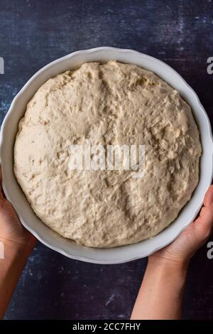 Mani che tengono la pasta turca di pane piatto dopo il primo aumento in a. recipiente grande su sfondo scuro Foto Stock