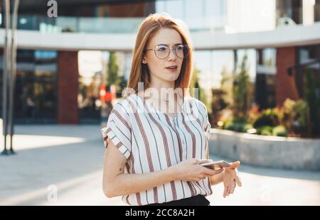 Donna caucasica confusa con capelli rossi e lenticelle che indossano occhiali posa fuori in strada tenendo un cellulare e guardando via Foto Stock