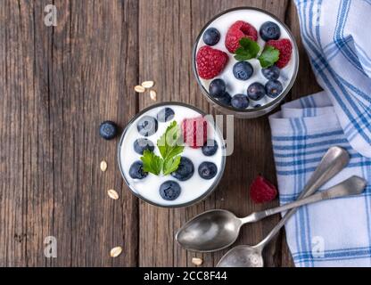 Yogurt bianco in due ciotole con mirtilli e lamponi su scrivania in legno naturale, vista piatta. Foto Stock