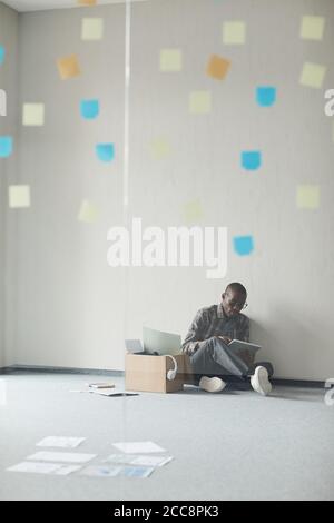 Uomo africano seduto sul pavimento e lavorando online utilizzando tablet digitale in una stanza vuota Foto Stock