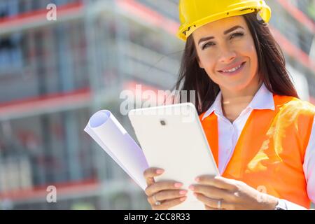 Donna sorridente in PPE tiene un tablet e un progetto con un cantiere in background. Foto Stock
