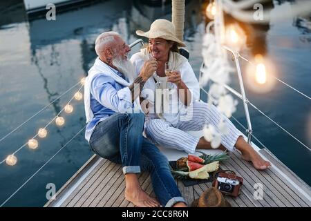 Coppia senior che brindano champagne e mangiano frutta in vacanza in barca a vela - persone anziane felici che si divertono a celebrare l'anniversario di matrimonio gita in barca Foto Stock