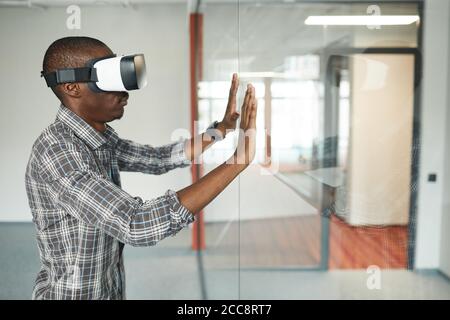 Vista laterale dell'uomo africano in realtà virtuale che tocca gli occhiali la parete di vetro durante il gioco di realtà virtuale nella stanza Foto Stock