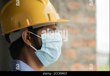 Vista laterale della testa del lavoratore addetto alla costruzione che vede all'esterno Costruzione della finestra di costruzione in Sad - lavoratore industriale in un hardhat con maschera medica Foto Stock