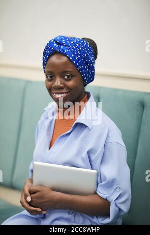 Ritratto di giovane donna africana seduta su divano con digitale tablet e sorridendo con la fotocamera Foto Stock