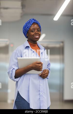 Ritratto di donna d'affari africana con sciarpa sulla sua presa della testa tablet pc e sorridendo con la fotocamera mentre si è in ufficio Foto Stock