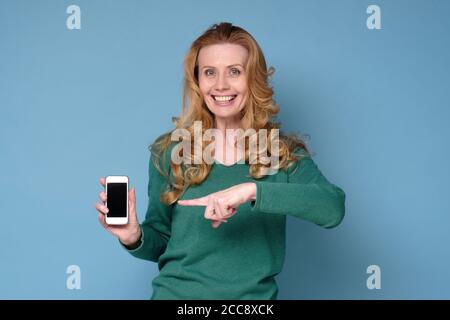 Sorridente donna bionda matura che tiene il telefono cellulare e punta ad esso Foto Stock