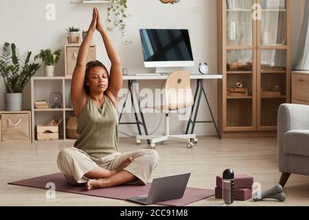 Giovane donna seduta sul tappetino da ginnastica di fronte al laptop e fare yoga a casa Foto Stock