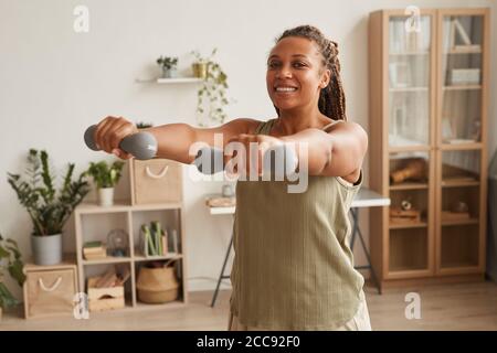 Giovane donna sorridente in piedi ed esercitandosi in manubri nella stanza a casa Foto Stock