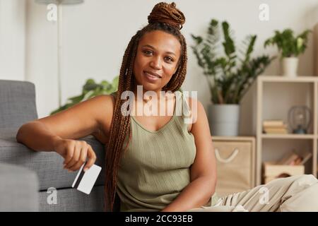 Ritratto di giovane donna africana con bella acconciatura sorridente fotocamera mentre usa la sua carta di credito a casa Foto Stock