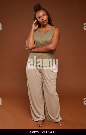 Ritratto di giovane donna africana con elegante acconciatura guardando fotocamera mentre si è in piedi contro lo sfondo marrone Foto Stock