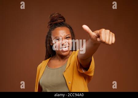 Ritratto di giovane donna africana sorridente e mostrante il pollice in su sulla fotocamera contro lo sfondo marrone Foto Stock