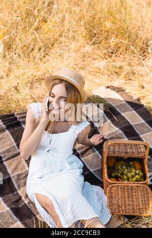 vista ad alto angolo della donna bionda in estate vestito che parla sullo smartphone vicino al cesto di vimini con frutta Foto Stock