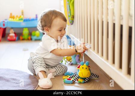 Sorridente bambina (16 mesi) seduta sul pavimento e giocando nella sua camera da letto con il suo giocattolo preferito coccolato vicino al suo presepe di legno bianco. Riproduzione ri Foto Stock