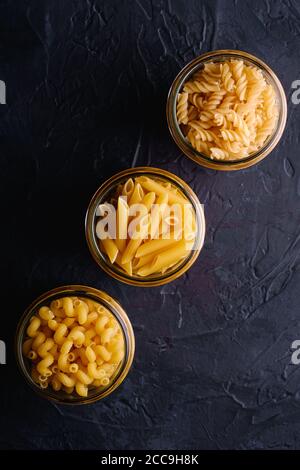 Tre vasetti di vetro in fila con una varietà di pasta di grano dorato non cotta su sfondo nero scuro testurizzato, vista dall'alto Foto Stock