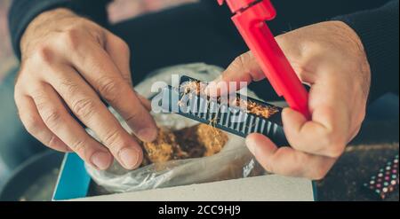 L'uomo fumatore arrotola sigarette di tabacco domestiche usando la macchina rotabile. Primo piano Foto Stock