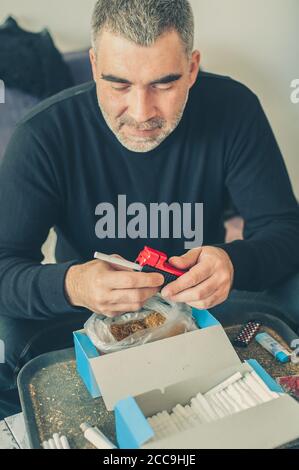 L'uomo fumatore arrotola sigarette di tabacco domestiche usando la macchina rotabile. Primo piano Foto Stock