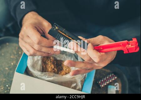 L'uomo fumatore arrotola sigarette di tabacco domestiche usando la macchina rotabile. Primo piano Foto Stock