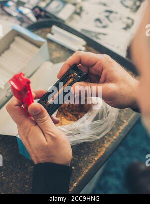 L'uomo fumatore arrotola sigarette di tabacco domestiche usando la macchina rotabile. Primo piano Foto Stock