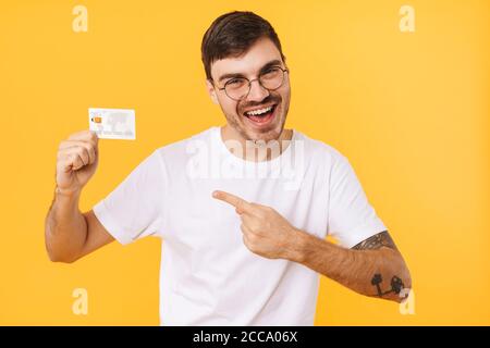 Foto di gioioso giovane uomo in occhiali che tiene e indica dito sulla carta di credito isolato su sfondo giallo Foto Stock