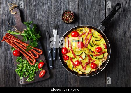 omelette con zucchine, salsicce affumicate sottili e pomodori in padella su un tavolo di legno scuro con ingredienti su un tagliere, vista orizzontale Foto Stock