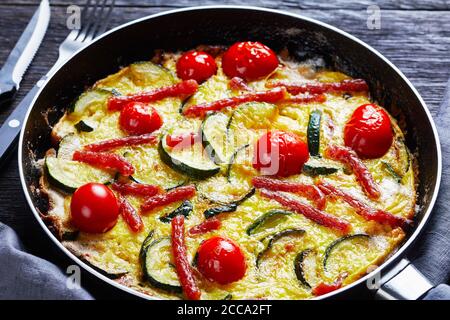 frittata, omelette con zucchine, sottili salsicce affumicate e pomodori ripieni in una padella su un tavolo di legno scuro con ingredienti sullo sfondo, atterrano Foto Stock