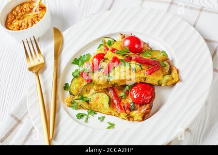 frittata, omelette con zucchine, sottili salsicce affumicate e pomodori ripieni su un piatto bianco su un tavolo di legno con ingredienti sullo sfondo, orizzonte Foto Stock