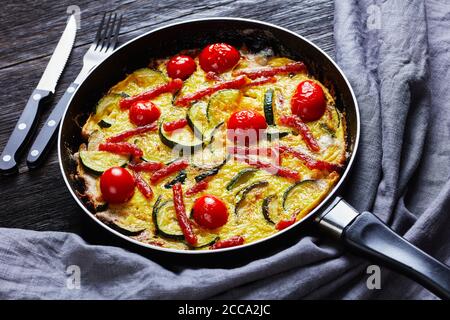 frittata, omelette con zucchine, sottili salsicce affumicate e pomodori ripieni in una padella su un tavolo di legno scuro, vista orizzontale dall'alto Foto Stock