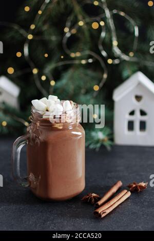 Cioccolata calda in un vasetto di vetro di muratore con marshmallows e cannella e spezie all'anice su tavola nera circondata da abeti, luci di Natale e decorazioni Foto Stock