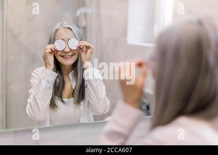 Cura della pelle del viso e procedure di bellezza. Ritratto di una donna con capelli grigi, divertente e anziana, in piedi nel bagno davanti allo specchio e alla copertura Foto Stock