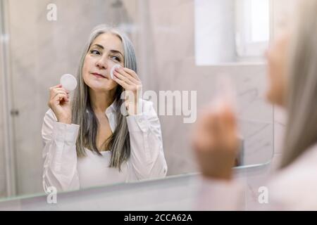 Concetto di igiene e cura della pelle. Affascinante signora grigio capelli senior utilizzando un tampone di cotone con acqua micellare per rimuovere il make up dal viso, in piedi Foto Stock