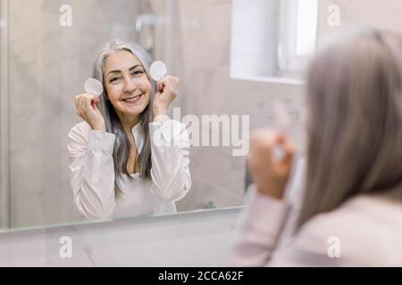 Sorridente donna abbastanza matura con bei capelli lunghi grigi, eseguendo la sua routine di cura della pelle, in posa in bagno luce di casa di fronte allo specchio con Foto Stock