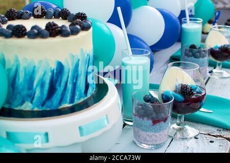 Tavolo dolce per feste per bambini. Decorate con palline blu e bianche. Torta decorata con mirtilli freschi e more, cocktail di latte in un gla alto Foto Stock