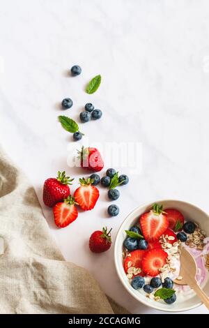 Colazione estiva. Ciotola di yogurt con fragole, mirtilli e muesli su uno sfondo di marmo bianco con lino beige e più frutta. Foto Stock